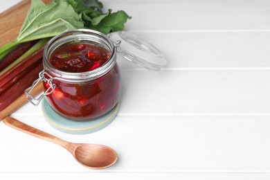 Photo of Jar of tasty rhubarb jam, stems and spoon on white wooden table. Space for text