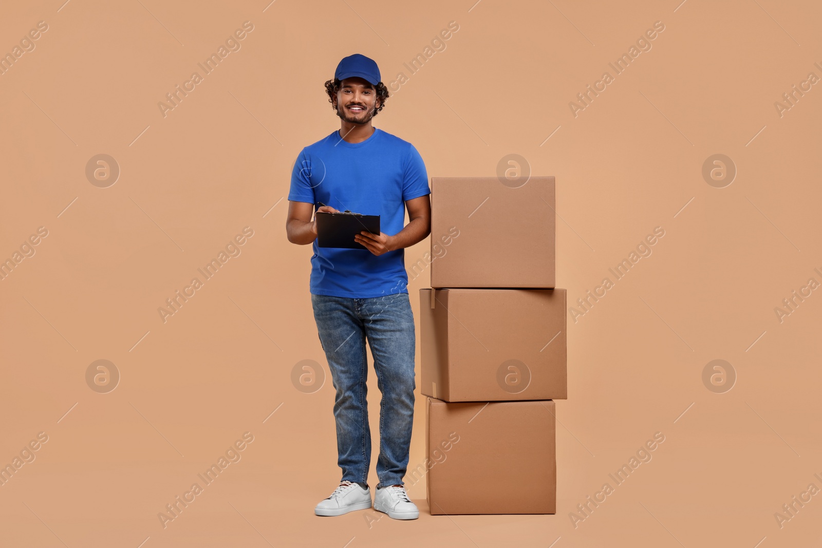 Photo of Happy courier with clipboard and stack of parcels on light brown background