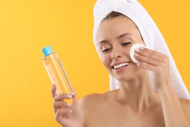 Smiling woman removing makeup with cotton pad and holding bottle on yellow background