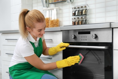 Photo of Female janitor cleaning oven with sponge in kitchen