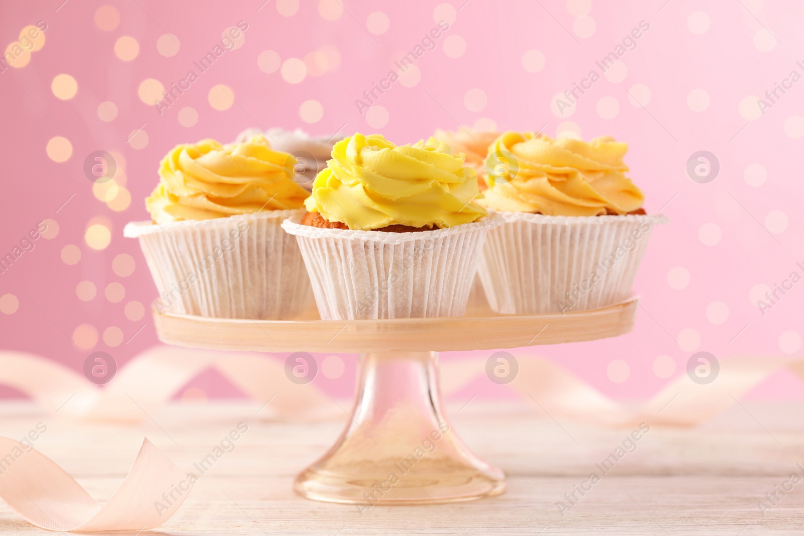 Photo of Stand with tasty cupcakes on white wooden table against blurred lights