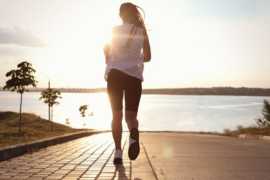 Photo of Young woman running near river in morning, back view