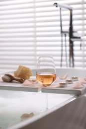 Photo of Wooden tray with wine, toiletries and flower petals on bathtub in bathroom