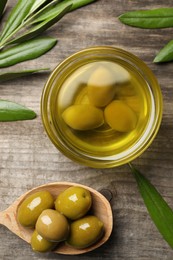 Photo of Cooking oil in bowl, olives and leaves on wooden table, flat lay