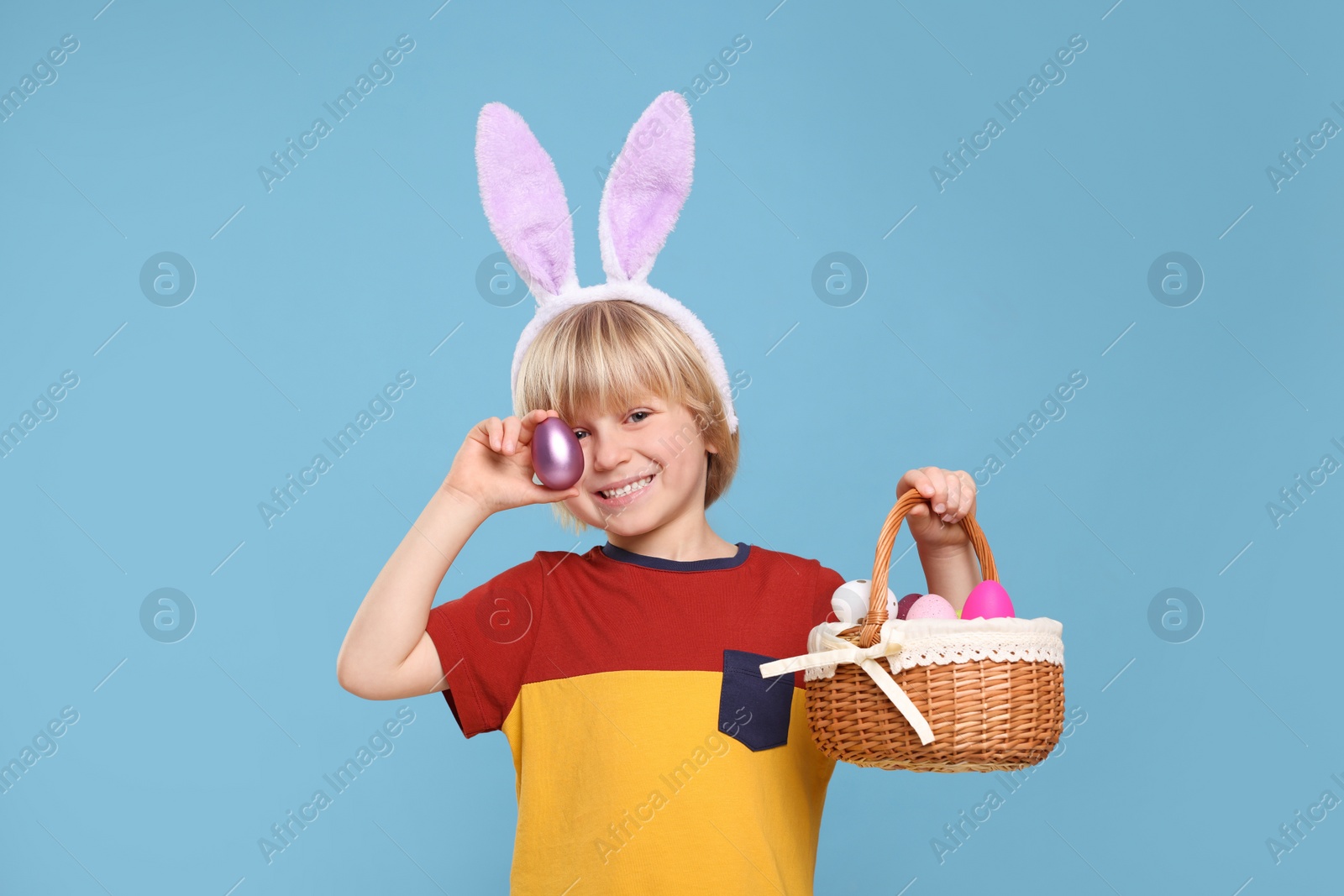 Photo of Happy boy in bunny ears headband holding wicker basket with painted Easter eggs on turquoise background