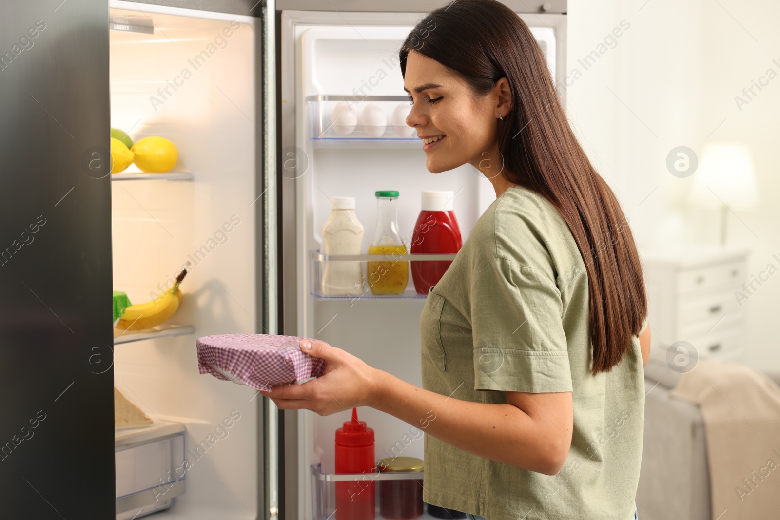 Photo of Happy woman holding bowl covered with beeswax food wrap near refrigerator in kitchen