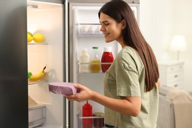 Happy woman holding bowl covered with beeswax food wrap near refrigerator in kitchen