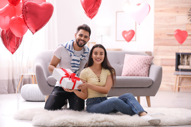Photo of Lovely young couple with gift box in living room decorated with heart shaped balloons. Valentine's day celebration