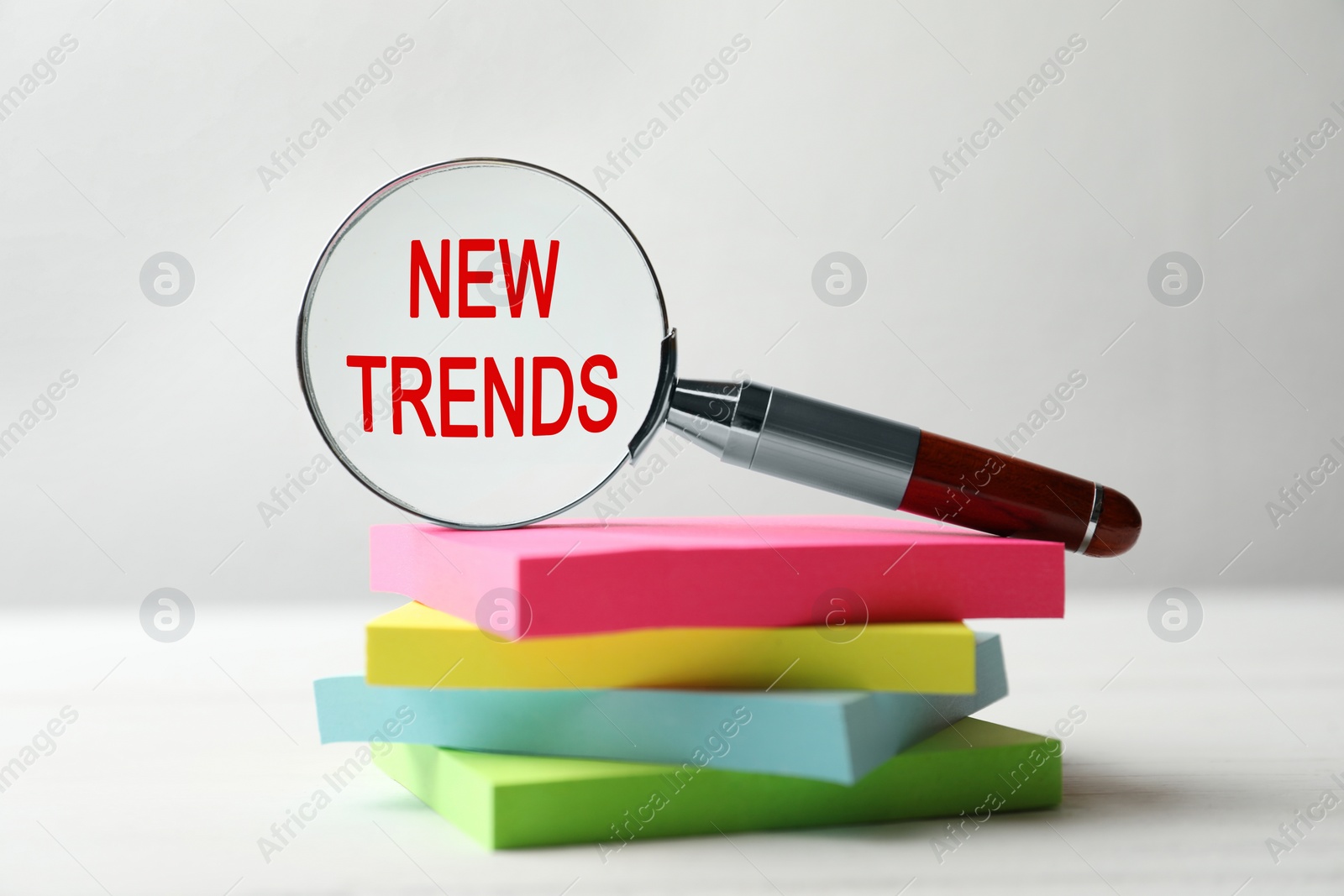 Image of Searching new and popular trends. Magnifier glass with words and stack of colorful stickers on white table, closeup