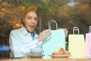 Photo of Special Promotion. Emotional young woman with shopping bags in cafe, view from outdoors