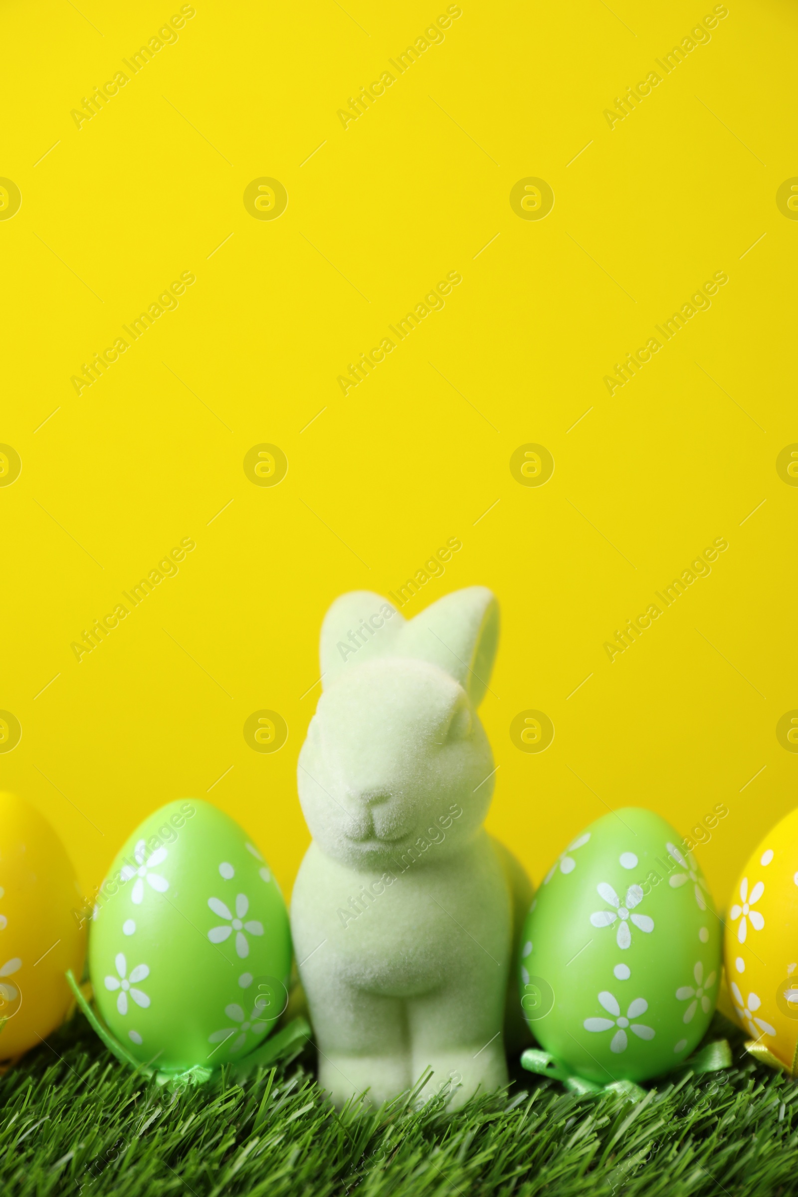 Photo of Easter bunny toy and dyed eggs on green grass against yellow background. Space for text