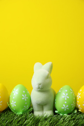 Photo of Easter bunny toy and dyed eggs on green grass against yellow background. Space for text