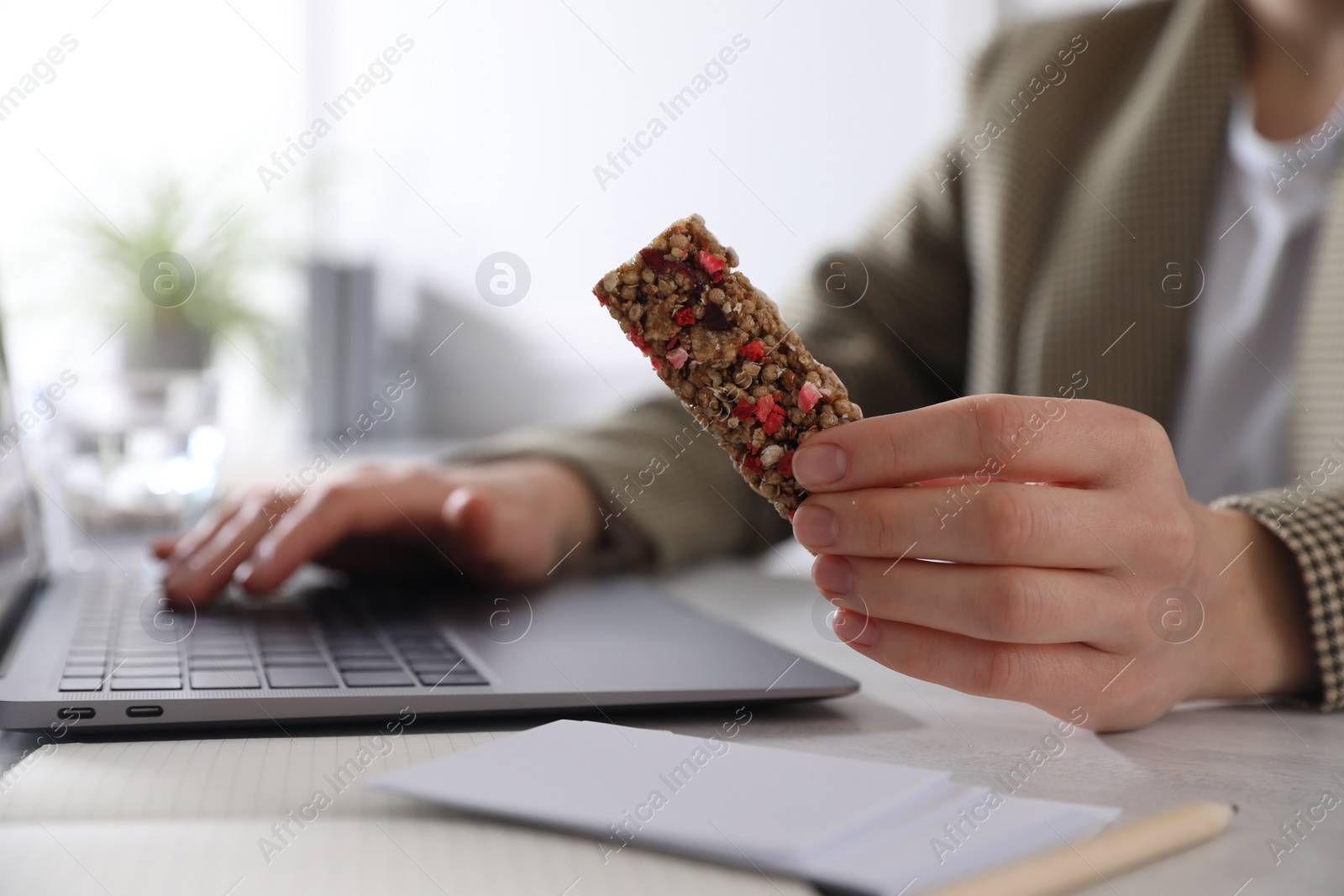 Photo of Woman holding tasty granola bar working with laptop at light table in office, closeup