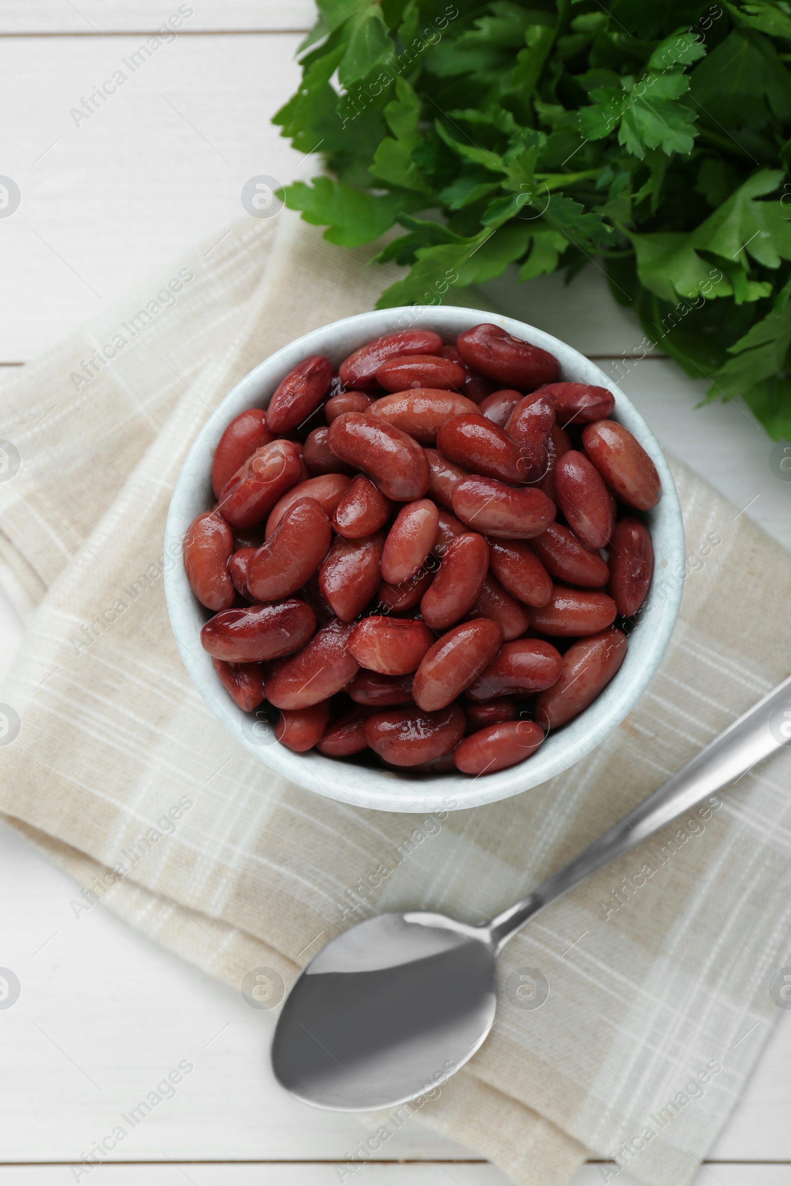 Photo of Bowl of canned kidney beans, parsley and spoon on white wooden table, top view