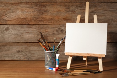 Easel with blank canvas board and painting tools for children on table near wooden wall. Space for text