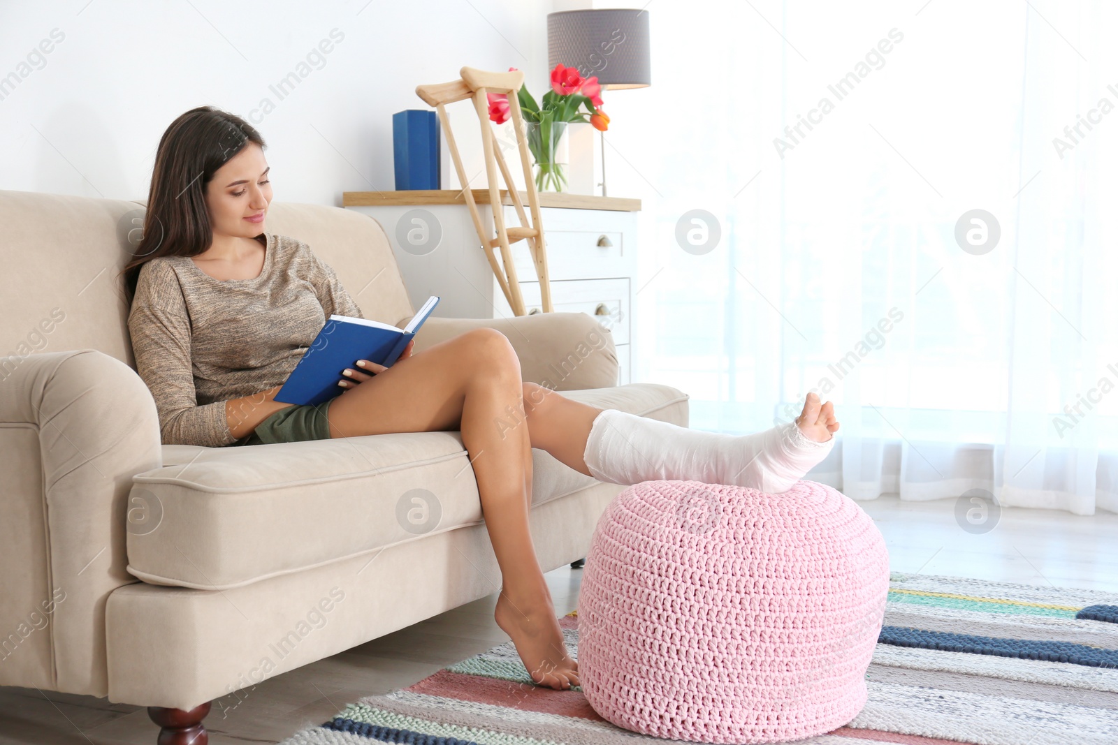 Photo of Young woman with broken leg in cast reading book while sitting on sofa at home