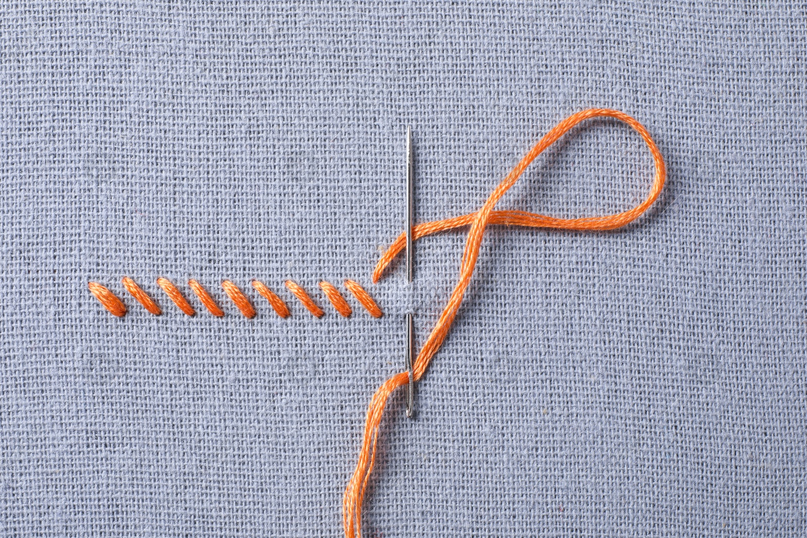 Photo of Needle with orange embroidery floss and row of stitches on grey fabric, top view