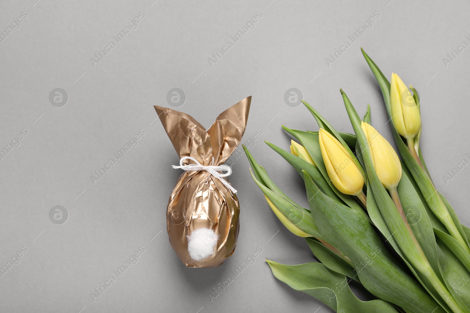 Photo of Easter bunny made of shiny gold paper and egg near beautiful tulips on grey background, flat lay. Space for text