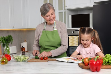 Cute little girl with her granny cooking by recipe book in kitchen