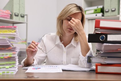 Photo of Overwhelmed woman sitting at table with stacks of documents and folders in office