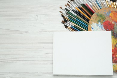 Photo of Blank canvas, brushes and palette on white wooden table, flat lay. Space for text