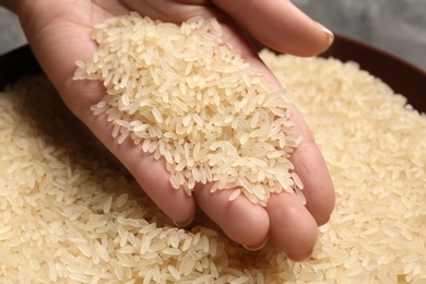 Photo of Woman holding grains near plate with parboiled rice on table, closeup