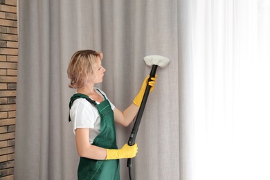 Female janitor removing dust from curtain with steam cleaner indoors
