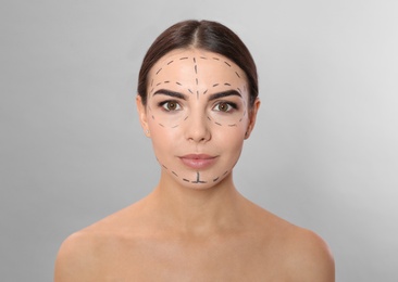 Young woman with marks on face for cosmetic surgery operation against grey background