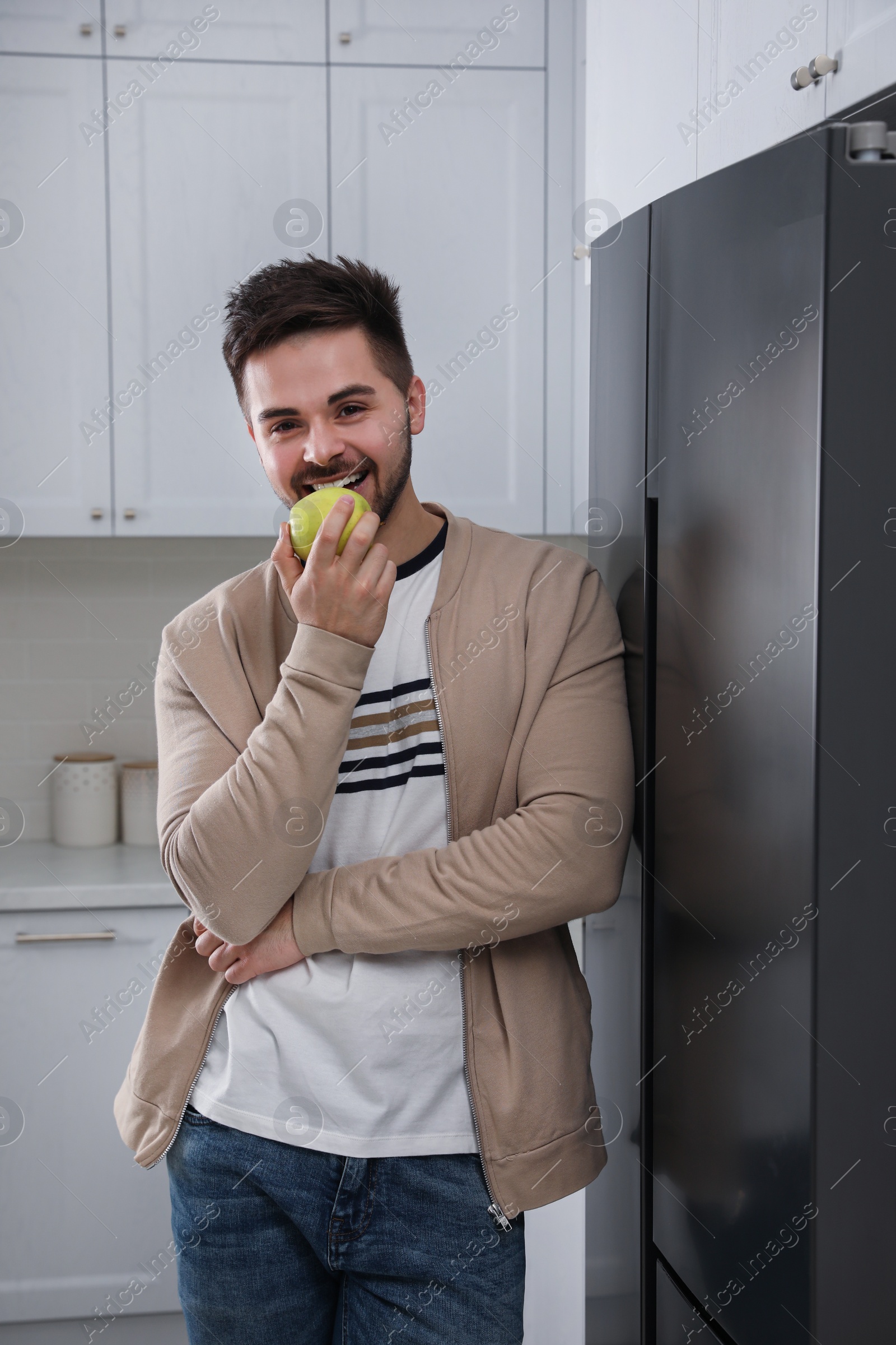 Photo of Young man eating apple near refrigerator in kitchen