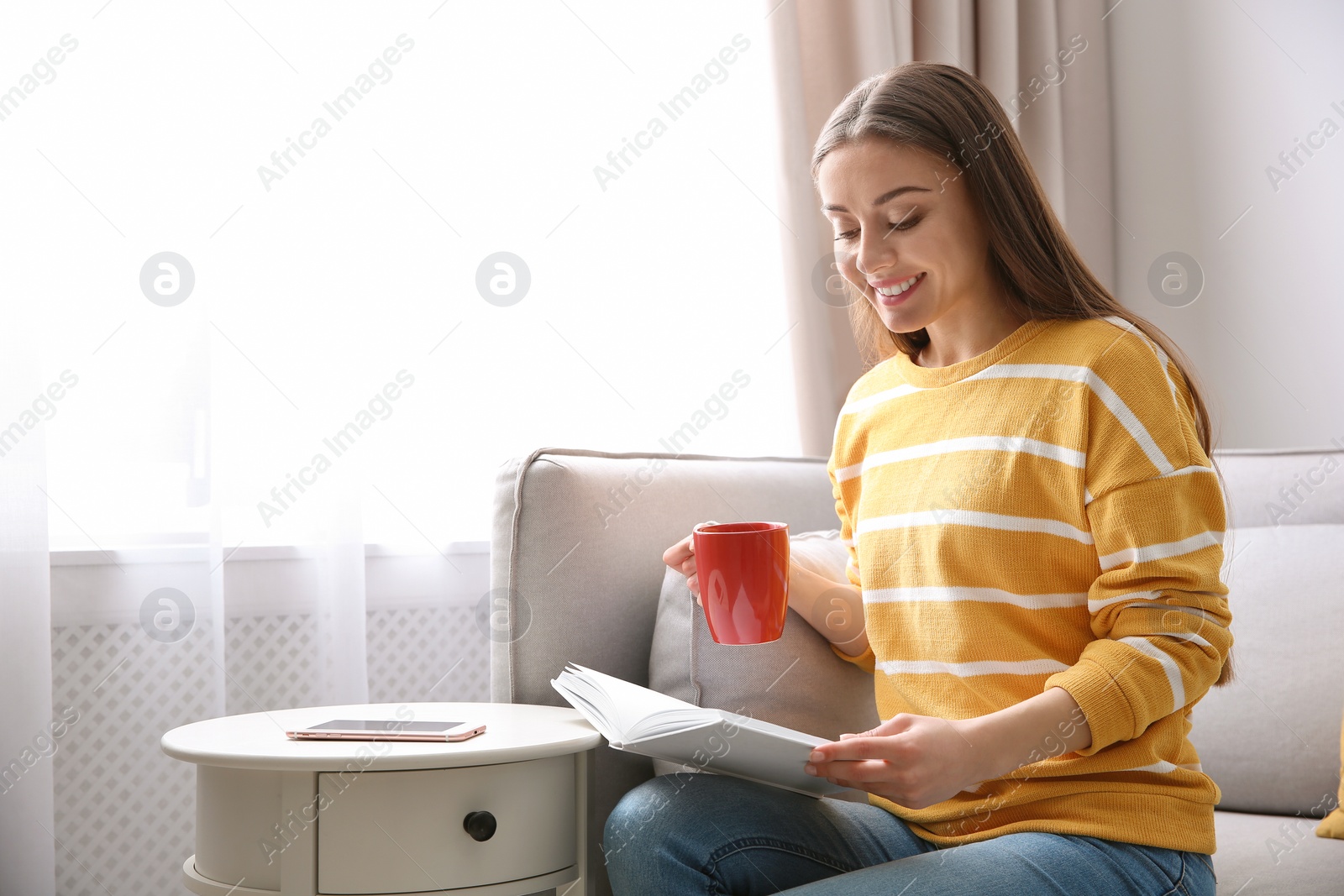 Photo of Young woman with cup of coffee reading book on couch at home