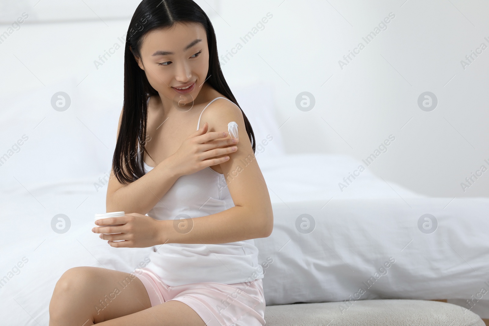 Photo of Beautiful young Asian woman applying body cream on shoulder in bedroom, space for text