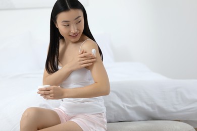 Beautiful young Asian woman applying body cream on shoulder in bedroom, space for text