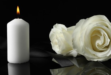 Photo of Burning candle, white roses and ribbon on black mirror surface in darkness, closeup. Funeral symbols