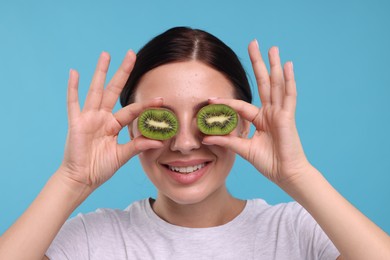 Photo of Young woman holding halves of kiwi near her eyes on light blue background