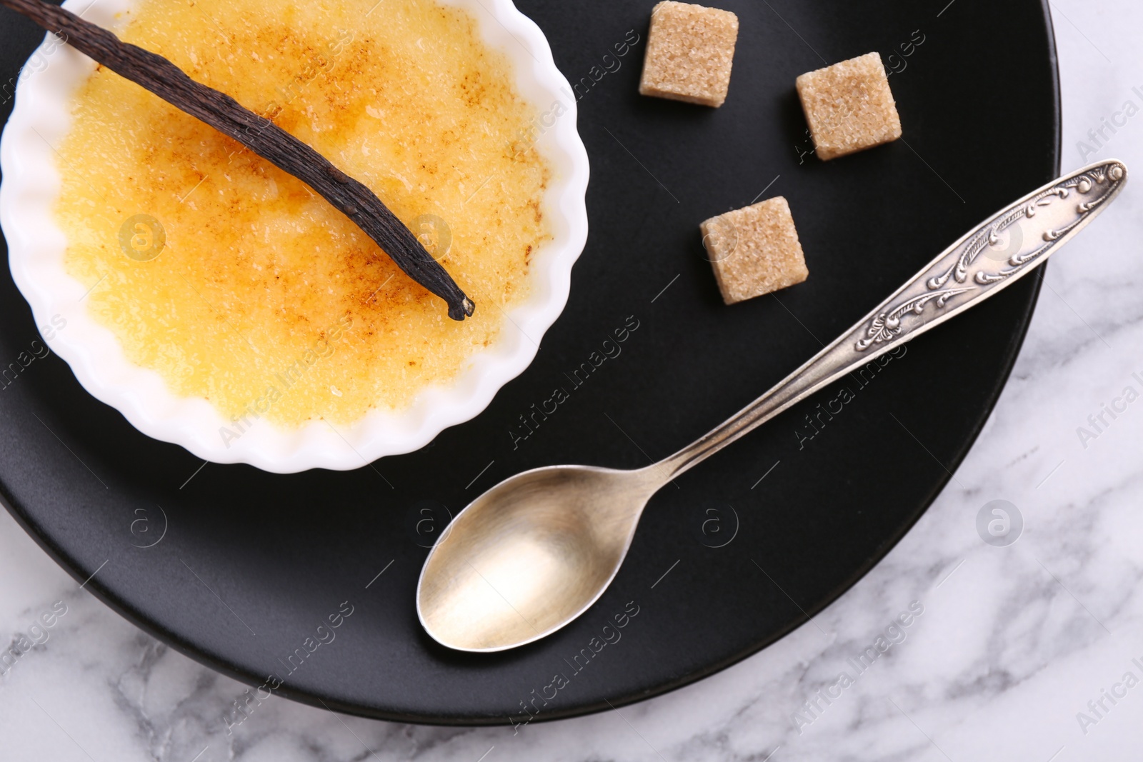 Photo of Delicious creme brulee in bowl, vanilla pod, sugar cubes and spoon on white marble table, top view