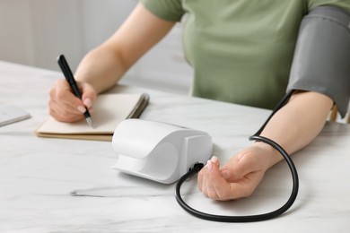 Photo of Woman measuring blood pressure and writing it down into notebook in kitchen, closeup