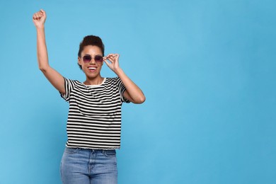 Happy young woman in stylish sunglasses dancing on light blue background. Space for text