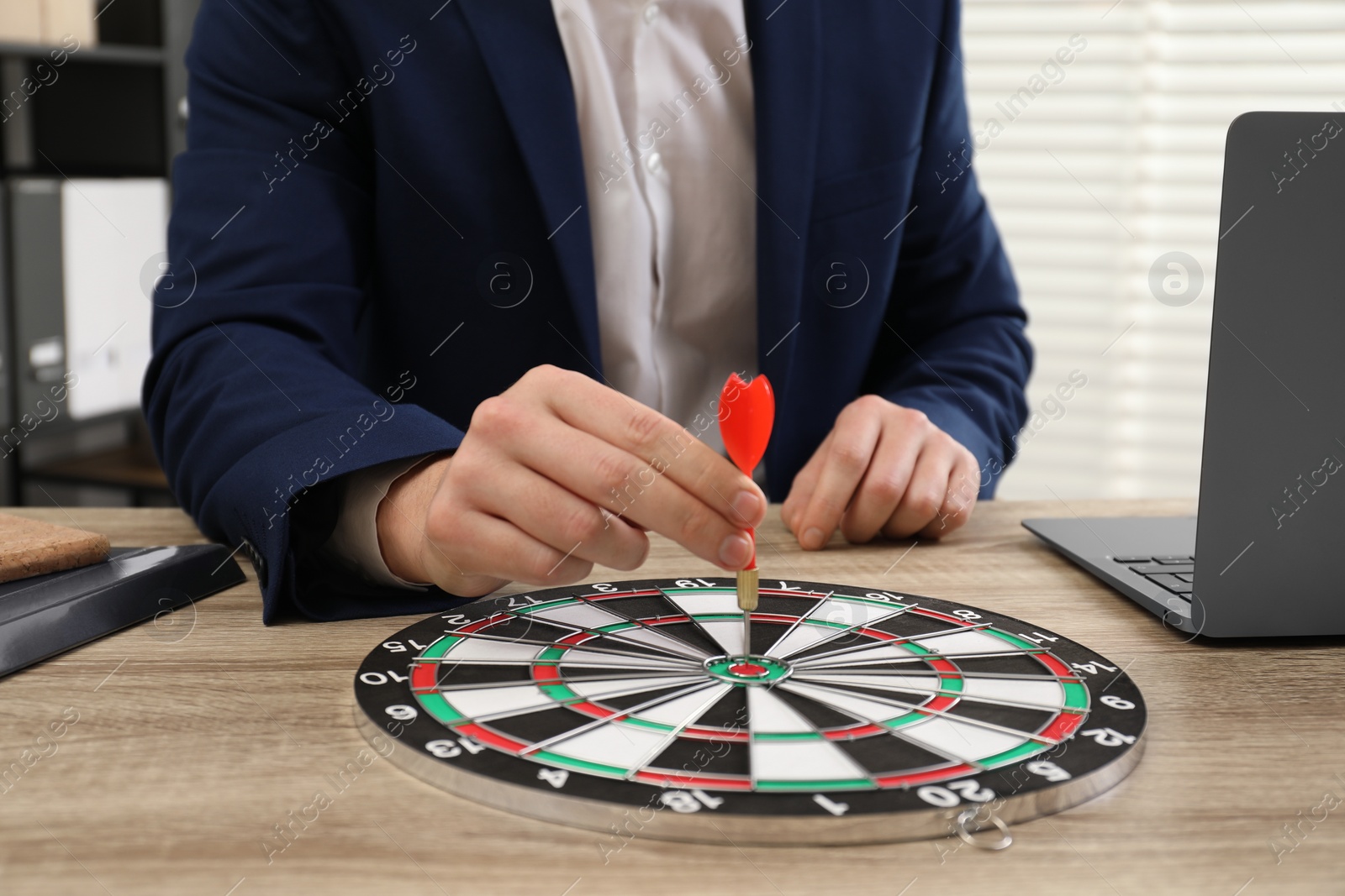 Photo of Business targeting concept. Man with dart aiming at dartboard at wooden table indoors, closeup