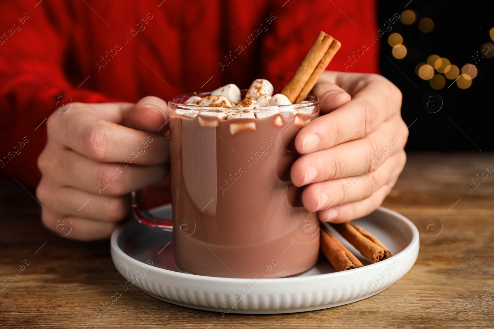 Photo of Woman holding glass cup of hot cocoa with aromatic cinnamon and marshmallows at wooden table against blurred lights, closeup