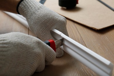 Photo of Worker cutting foam crown molding with utility knife at wooden table, closeup