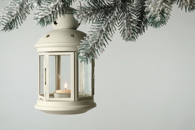 Photo of Christmas lantern with candle hanging on snowy fir tree branch against light background. Space for text