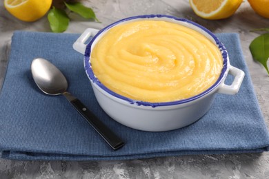 Photo of Delicious lemon curd in bowl, fresh citrus fruit, green leaves and spoon on grey table