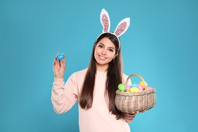 Photo of Beautiful woman in bunny ears headband holding basket with Easter eggs on color background