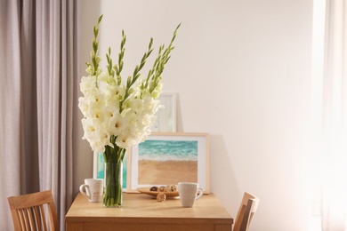 Vase with beautiful white gladiolus flowers, pictures and cups on wooden table in room, space for text