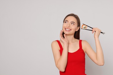 Photo of Happy young woman holding sushi roll with chopsticks on light background. Space for text