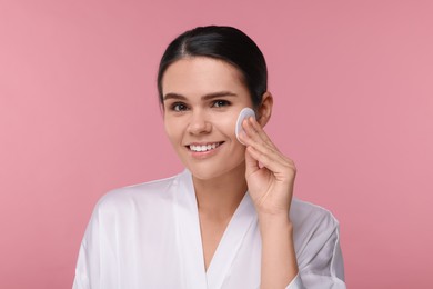 Young woman cleaning her face with cotton pad on pink background