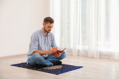 Photo of Muslim man reading Koran on prayer rug indoors