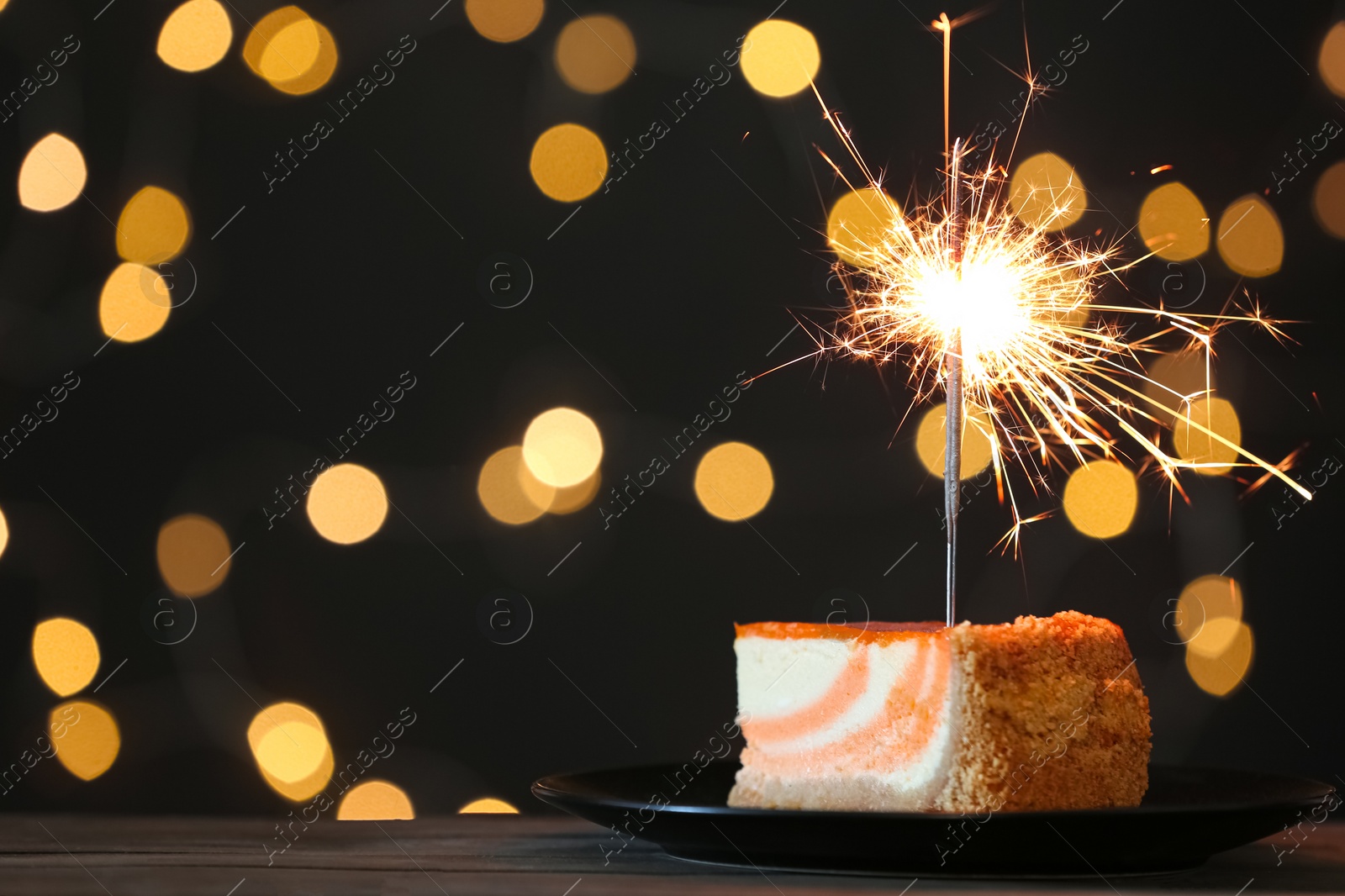 Photo of Cake with burning sparkler on black table against blurred festive lights. Space for text