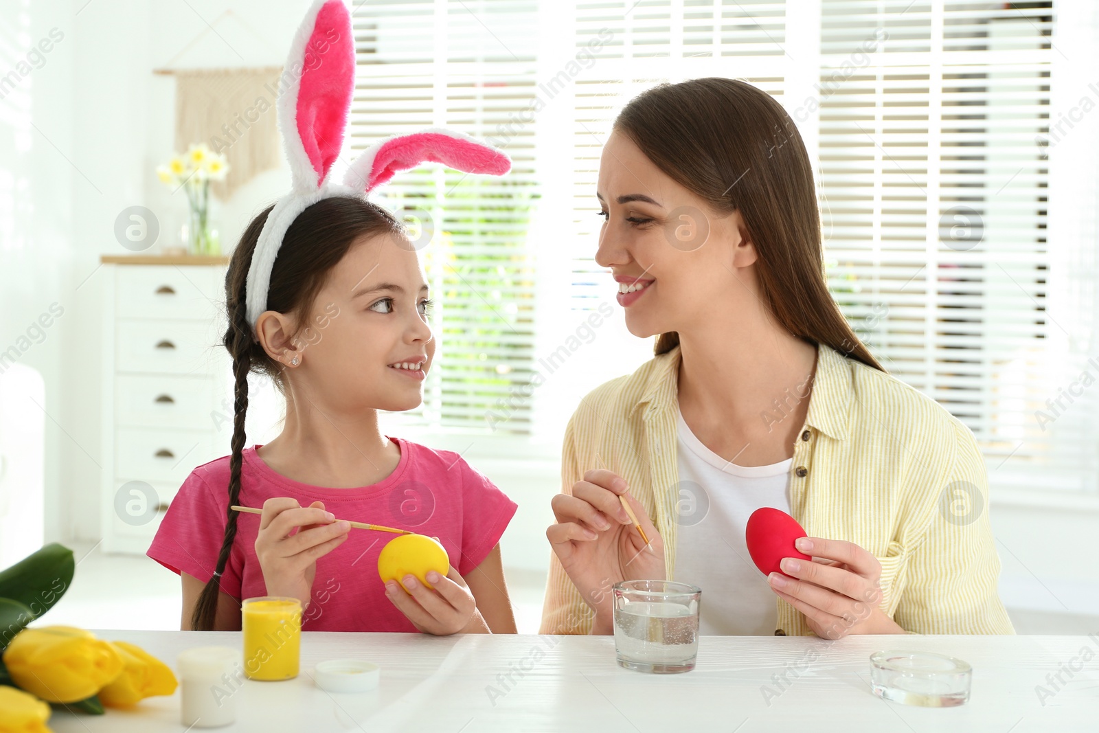 Photo of Happy daughter with bunny ears headband and her mother painting Easter eggs at home