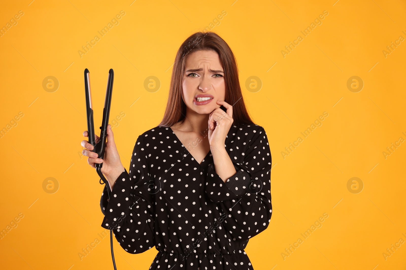 Photo of Upset young woman with flattening iron on yellow background. Hair damage